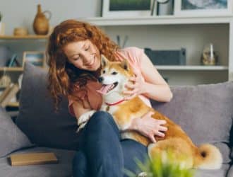 A red-haired woman sitting on a living room couch as she cuddles with a happy shiba inu that’s half sitting in her lap.