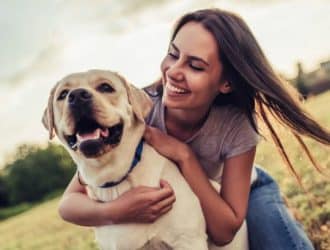 A young woman in a purple shirt and jeans hugs her dog in a park. She and the dog are smiling and happy.