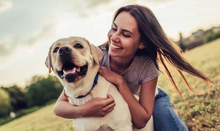 A young woman in a purple shirt and jeans hugs her dog in a park. She and the dog are smiling and happy.