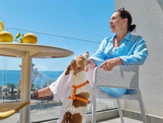 A woman sitting on her balcony on a sunny day and laughing as her small dog stands up for her attention.