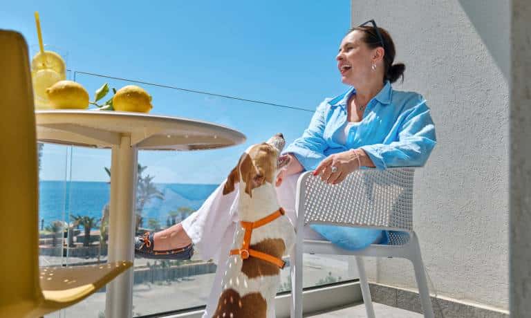 A woman sitting on her balcony on a sunny day and laughing as her small dog stands up for her attention.