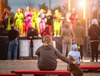 A dog owner sitting on an outdoor bench with her dog. They are at an outdoor festival watching a show on the mainstage.