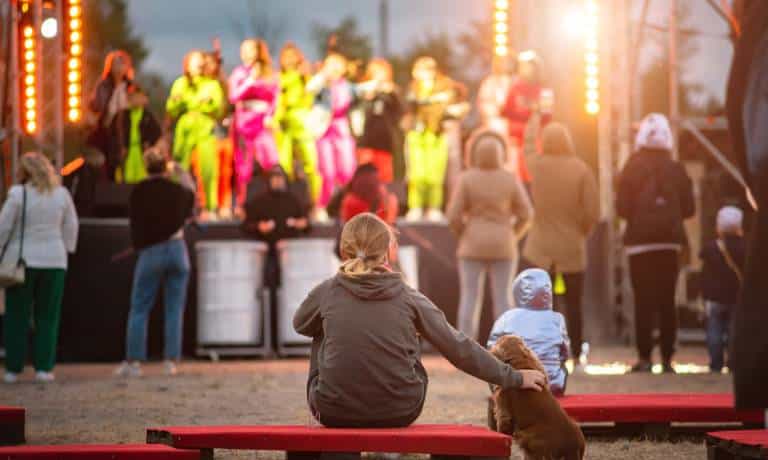 A dog owner sitting on an outdoor bench with her dog. They are at an outdoor festival watching a show on the mainstage.