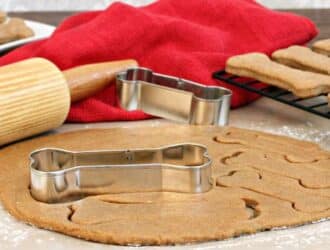 A red towel, a wooden rolling pin, a cooling rack with cooked treats, and dog bone-shaped cookie cutters sit on a counter.