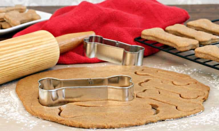 A red towel, a wooden rolling pin, a cooling rack with cooked treats, and dog bone-shaped cookie cutters sit on a counter.