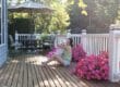 A woman sits on a wooden deck hugging a little white dog. Patio furniture and two pots with pink flowers appear on the deck.