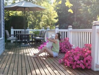 A woman sits on a wooden deck hugging a little white dog. Patio furniture and two pots with pink flowers appear on the deck.