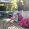 A woman sits on a wooden deck hugging a little white dog. Patio furniture and two pots with pink flowers appear on the deck.