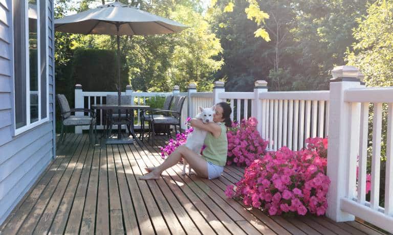 A woman sits on a wooden deck hugging a little white dog. Patio furniture and two pots with pink flowers appear on the deck.