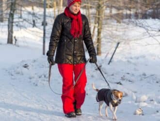 A woman wearing a winter coat, gloves, and a scarf is walking her dog through the snow. The dog is wearing a coat.