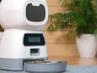 An unmarked white automatic pet food dispenser with a gray bowl sitting on the floor of a home.