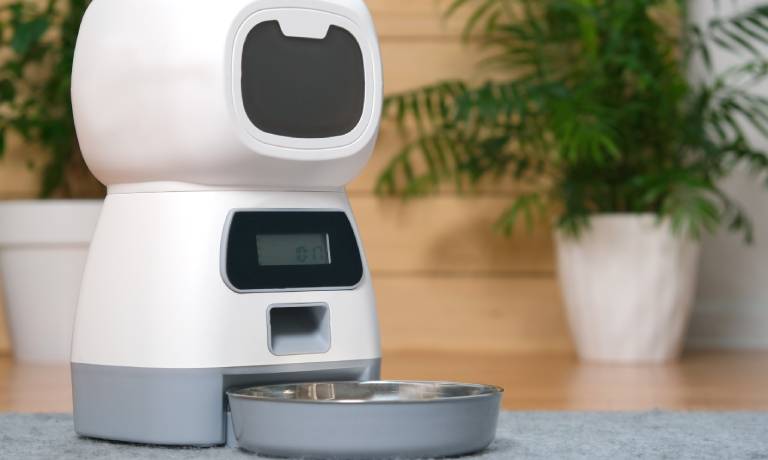 An unmarked white automatic pet food dispenser with a gray bowl sitting on the floor of a home.