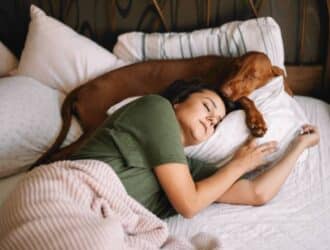 A woman in a green shirt and a brown dog sleeping in the same bed and using the same white pillow.