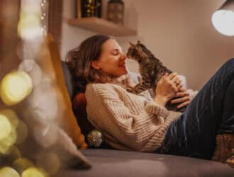 A woman siting on the couch as she cradles a brown tabby cat in a dimly-lit living room with fairly lights.