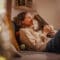 A woman siting on the couch as she cradles a brown tabby cat in a dimly-lit living room with fairly lights.