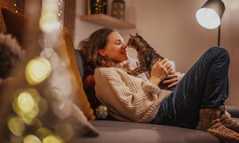 A woman siting on the couch as she cradles a brown tabby cat in a dimly-lit living room with fairly lights.